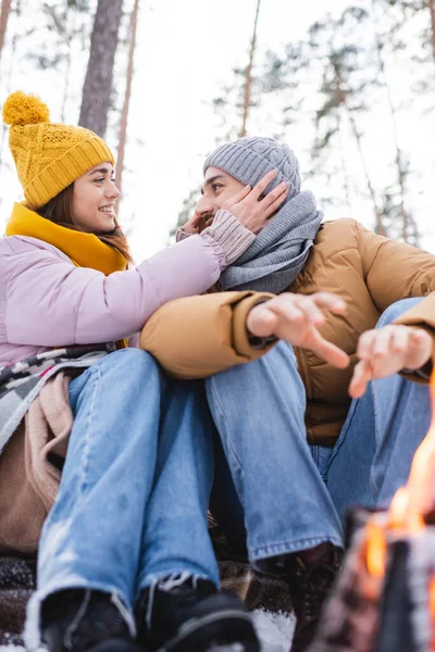 Tiefer Blickwinkel auf lächelnde Frau, die Strickmütze ihres Freundes in der Nähe des verschwommenen Lagerfeuers im Winterpark justiert — Stockfoto