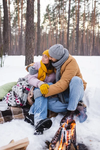 Mann umarmt Freundin in Decke neben Tassen und verschwommenem Lagerfeuer — Stockfoto