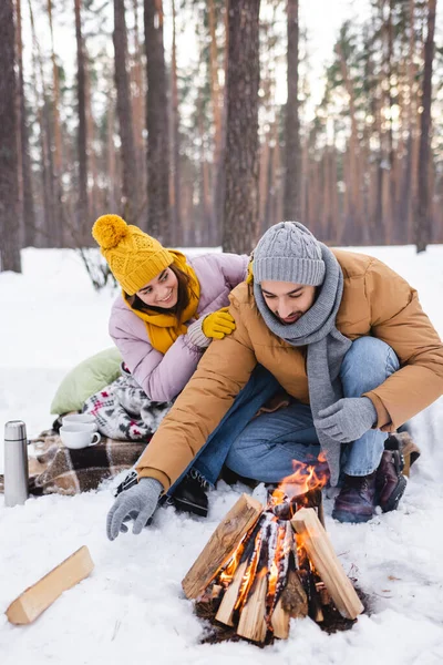 Lächelnde Frau umarmt Freund in der Nähe von Tassen, Thermoskannen und Lagerfeuer im Winterpark — Stockfoto