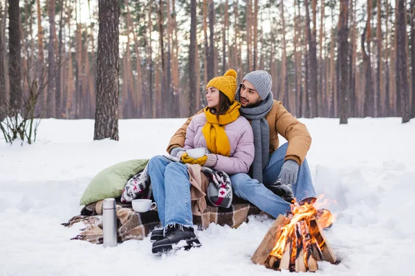 Jovem casal em roupa de inverno olhando para longe perto de copos e fogueira no parque de inverno — Fotografia de Stock