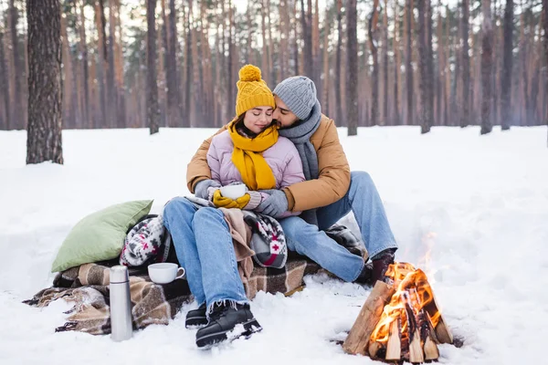 Mann küsst Freundin mit Tasse in der Nähe von Decken und Lagerfeuer im Winterpark — Stockfoto