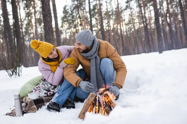 Lächelnde Frau mit Handschuhen am Lagerfeuer und Tassen im Winterwald — Stockfoto