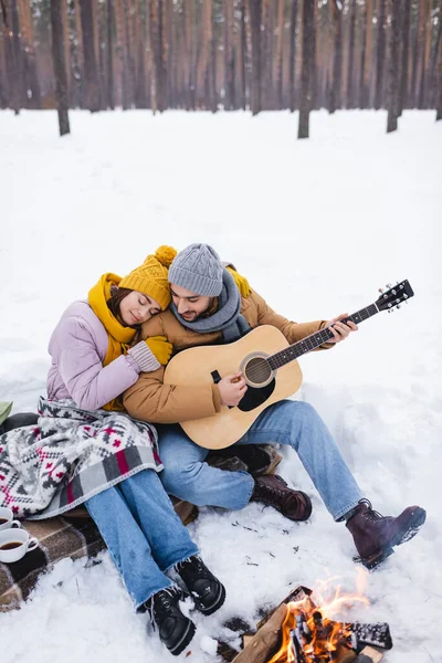 Lächelnder Mann spielt Akustikgitarre neben Freundin und Lagerfeuer im Winterpark — Stockfoto