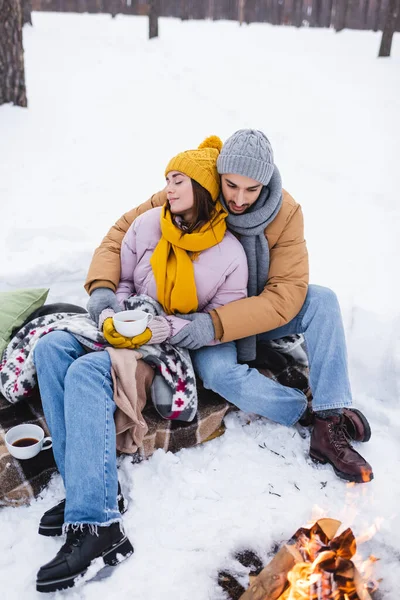 Mann umarmt Freundin mit Tasse Kaffee am Lagerfeuer im Winterpark — Stockfoto