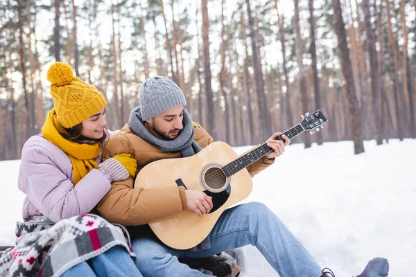 Lächelnde Frau umarmt Freund, der im Winterpark Akustikgitarre spielt — Stockfoto