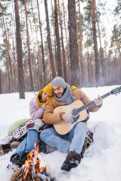 Mann spielt Akustikgitarre neben Freundin und verschwommenes Lagerfeuer im Winterpark — Stockfoto