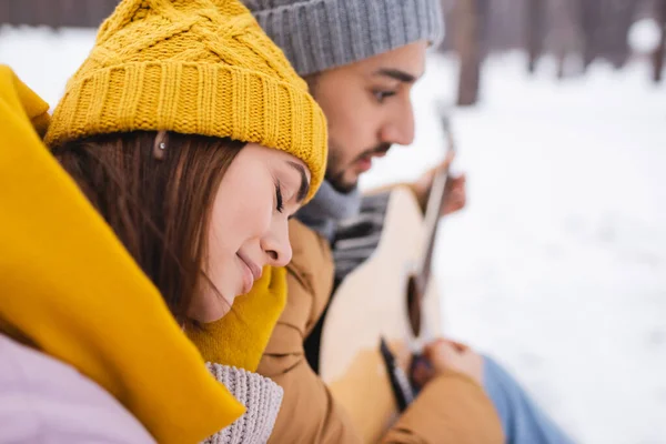 Side view of woman sitting near blurred boyfriend with acoustic guitar in winter park — Stock Photo