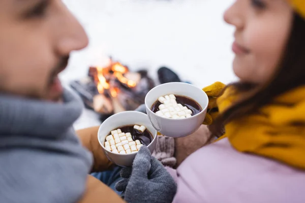 Coupé vue des tasses de cacao avec guimauves dans les mains du couple flou dans le parc d'hiver — Photo de stock