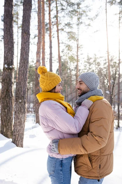 Вид сбоку на веселого мужчину, обнимающего девушку в вязаной шляпе и шарфе в зимнем парке — стоковое фото