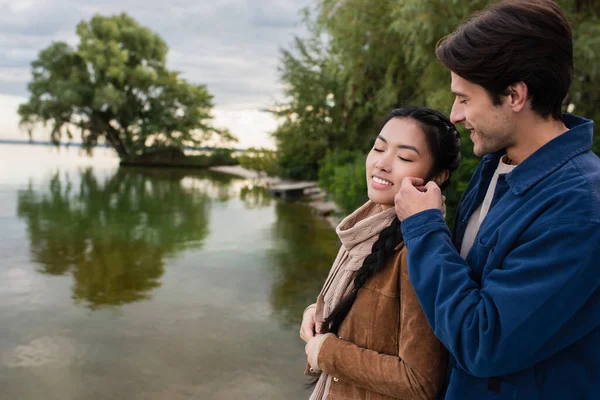 Homme souriant toucher la joue de petite amie asiatique près du lac — Photo de stock