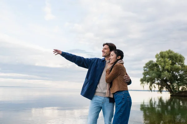 Smiling man hugging asian girlfriend and pointing with hand near lake — Stock Photo