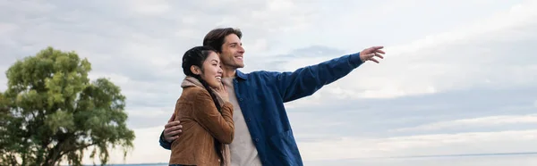 Cheerful man pointing with hand and hugging asian girlfriend with lake at background, banner — Stock Photo