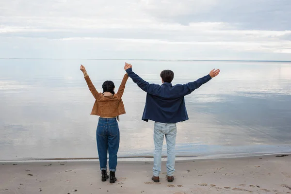 Vue arrière du jeune couple se tenant la main sur la plage près de la mer en automne — Photo de stock