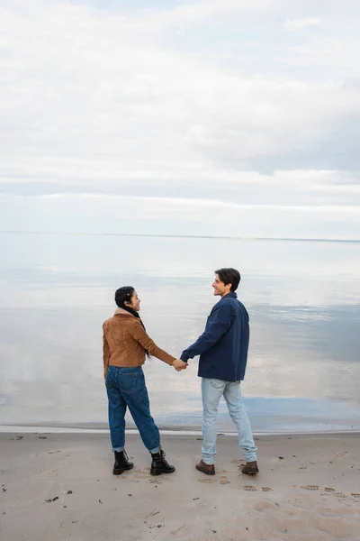 Vista laterale della coppia interrazziale sorridente che si tiene per mano vicino al mare durante il fine settimana — Foto stock
