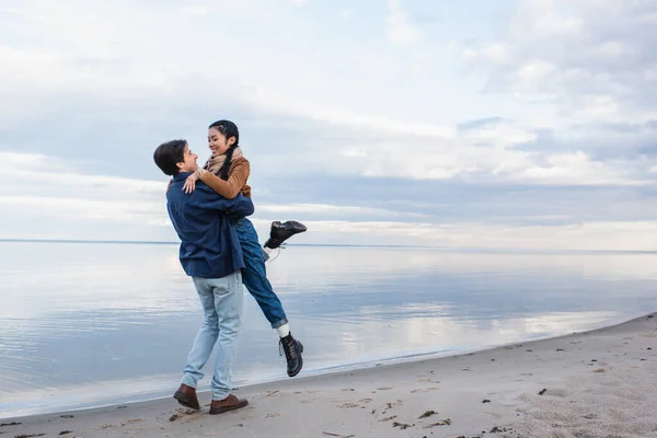 Man lifting asian girlfriend on beach near lake — Stock Photo