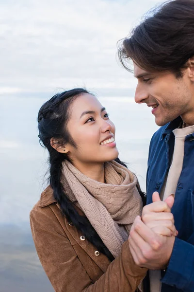 Sonriente pareja multiétnica tomados de la mano mientras se miran cerca del lago - foto de stock