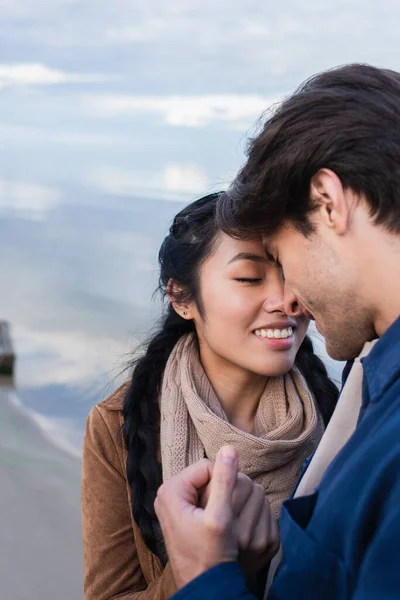 Smiling asian woman holding hand of boyfriend near blurred sea in autumn — Stock Photo