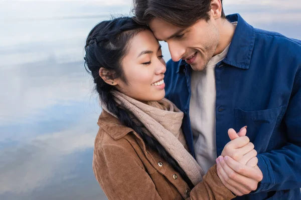 Glückliches multiethnisches Paar hält Händchen nahe verschwommenem Meer im Hintergrund — Stockfoto