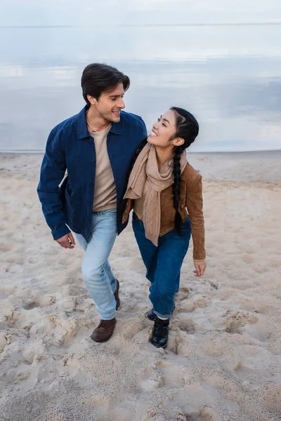 Cheerful interracial couple walking on beach near sea — Stock Photo