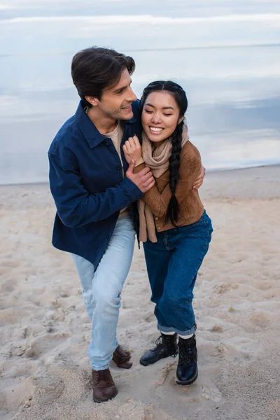 Мужчина обнимает весёлую азиатскую подружку на пляже — стоковое фото