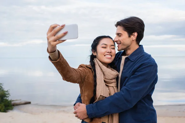 Улыбающийся мужчина обнимает азиатскую подружку и делает селфи на пляже. — стоковое фото