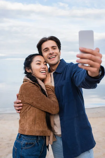 Sourire interracial couple câlin et prendre selfie sur flou smartphone sur la plage — Photo de stock