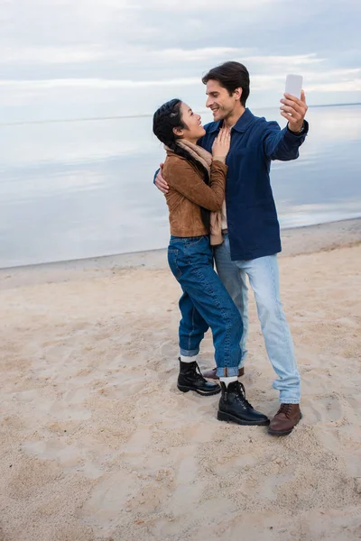 Vue latérale du couple interracial prenant selfie sur téléphone portable sur la plage en automne — Photo de stock