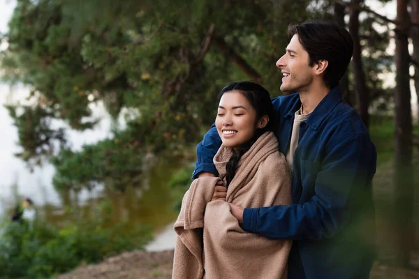 Smiling man hugging asian girlfriend in blanket outdoors — Stock Photo