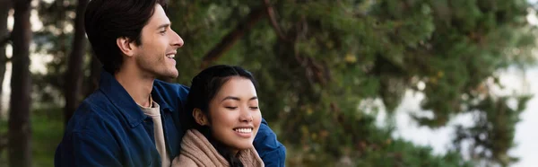 Sorrindo homem de pé perto asiático namorada com fechado olhos ao ar livre, banner — Fotografia de Stock