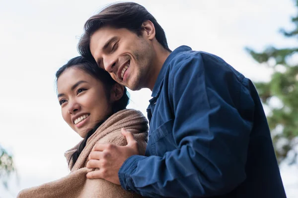 Bajo ángulo vista de hombre con los ojos cerrados abrazando asiático novia en manta al aire libre - foto de stock