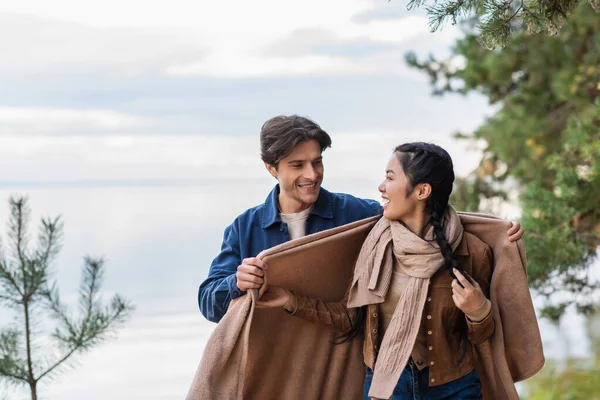 Man holding blanket near asian girlfriend with lake on background — Stock Photo