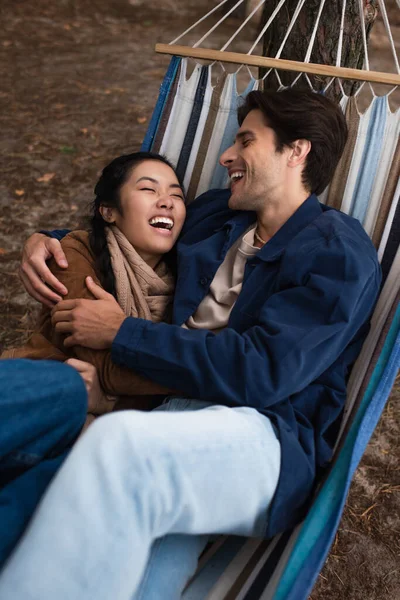 Hombre abrazando excitado asiático novia en hamaca al aire libre - foto de stock