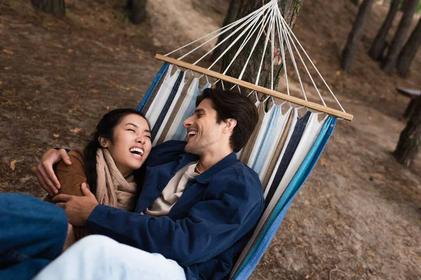 Emocionada pareja multiétnica abrazándose en hamaca durante el fin de semana - foto de stock