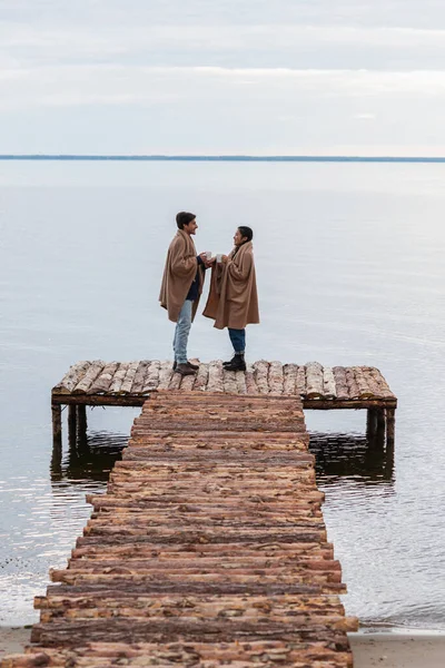 Vue latérale du couple interracial joyeux avec des tasses debout sur la jetée près de la mer — Photo de stock
