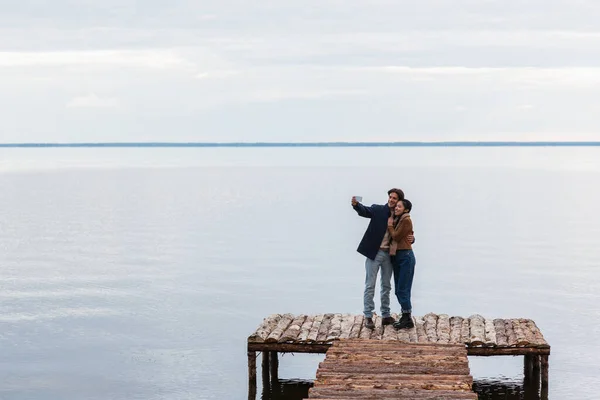 Feliz pareja multiétnica tomando selfie en el teléfono celular en el muelle cerca del mar — Stock Photo