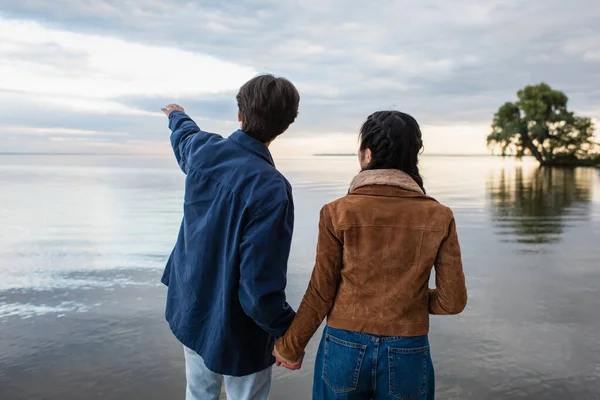 Vista trasera de la pareja cogida de la mano y el hombre apuntando al mar - foto de stock