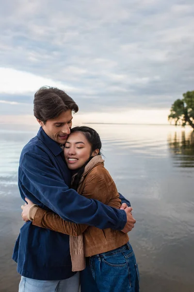 Sonriente hombre abrazando asiático novia con cerrado ojos cerca de mar - foto de stock