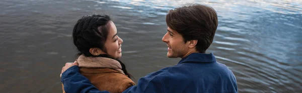 Vista lateral de la joven pareja multiétnica sonriéndose el uno al otro cerca del mar, pancarta - foto de stock