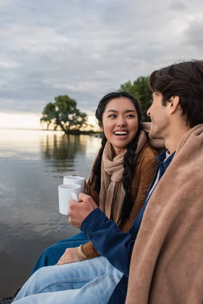Улыбающаяся азиатка держит кубок рядом с парнем и озером — стоковое фото