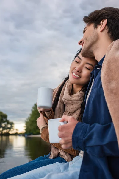 Man holding cup near smiling asian woman and lake during weekend — Stock Photo