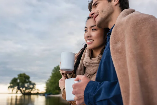 Smiling multiethnic couple in blanket holding cups near blurred lake — Stock Photo