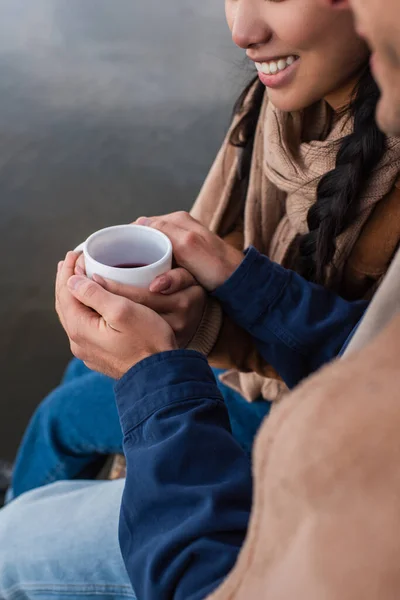 Обрезанный вид человека, держащегося за руки улыбающейся девушки с чашкой возле размытого озера — стоковое фото