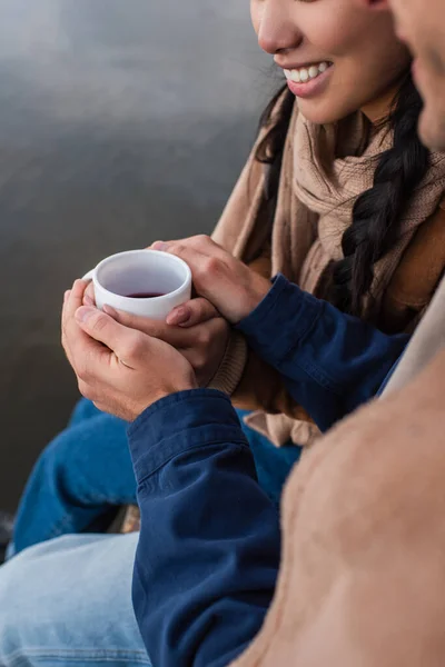 Vue recadrée de l'homme tenant la main d'une petite amie souriante avec tasse près du lac flou — Photo de stock