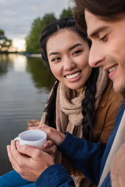 Усміхнена азіатка тримає чашку біля розмитого хлопця і озера на відкритому повітрі — стокове фото