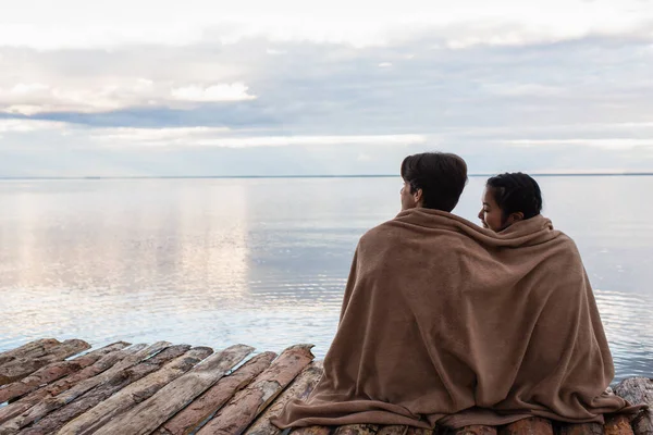 Межрасовая пара в одеяле сидит на деревянном пирсе у моря — стоковое фото