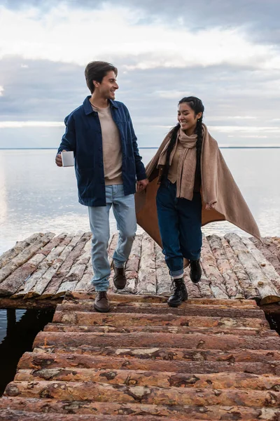 Sorridente coppia interrazziale con coperta e tazza a piedi sul molo vicino al mare — Foto stock