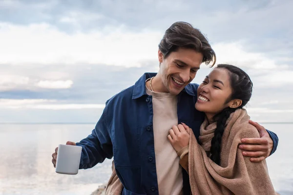 Happy asian woman in blanket embracing boyfriend with cup near lake during weekend — Stock Photo