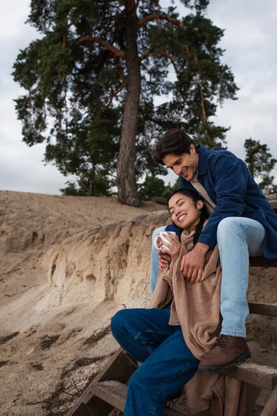 Uomo abbracciando donna asiatica con tazza e coperta sulle scale vicino collina — Foto stock