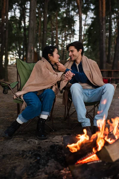 Vue latérale du couple interracial souriant assis sur des chaises près du feu de camp à l'extérieur — Photo de stock