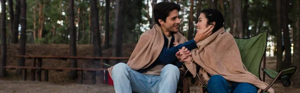 Smiling man touching face of asian girlfriend on chairs outdoors, banner — Stock Photo